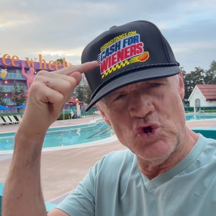 NASCAR legend Kenny Wallace pointing to his Cash for Wieners hat